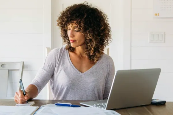 A woman with curly brown hair sits at a wooden table in a brightly lit room, writing in a small notebook while looking at papers spread out next to a silver laptop.